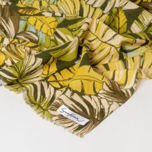 Un tissu léger avec un motif de feuilles tropicales vertes, jaunes et beiges, partiellement plié, avec une petite étiquette Soykan sur le coin.