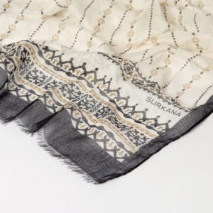 Écharpe à motifs crème et noirs, avec des dessins géométriques et floraux, des franges et le nom de la marque SURKANA imprimé dans un coin.