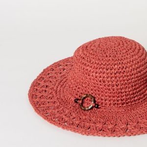 Chapeau de paille tressée rouge à large bord, doté d'une bande décorative et d'une boucle ronde en écaille, présenté sur un fond blanc uni.