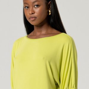 Femme portant un haut ample vert clair avec des manches au coude et des boucles d'oreilles dorées, debout sur un fond uni.