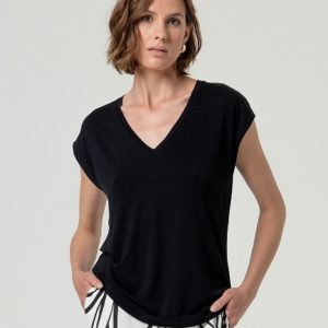 Une femme aux cheveux bruns courts porte un T-shirt noir à col en V et un pantalon blanc à motifs. Elle se tient debout, les mains dans les poches, sur un fond uni.