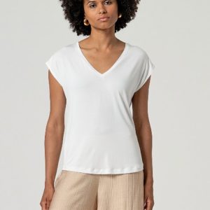 Une personne se tient debout, de face, vêtue d'un haut blanc à col en V et à manches courtes et d'un pantalon beige à rayures, sur un fond clair uni.