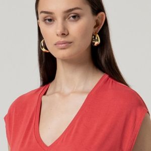 Femme aux longs cheveux bruns portant un haut rouge à col en V et de grandes boucles d'oreilles dorées, posant sur un fond clair uni.