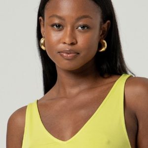 Une femme aux cheveux noirs et raides, portant des boucles d'oreilles en or et un haut jaune sans manches, regarde l'appareil photo sur un fond uni.