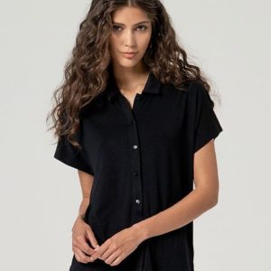Une femme aux longs cheveux bruns ondulés porte une chemise noire boutonnée à manches courtes et un pantalon de couleur claire, debout sur un fond clair uni.