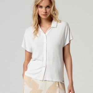Une femme aux cheveux blonds ondulés porte un chemisier blanc à manches courtes et une jupe beige à motif de feuilles, debout sur un fond clair uni.