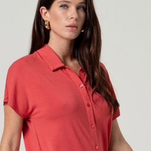 Une femme aux longs cheveux bruns porte une chemise boutonnée à manches courtes de couleur corail et des boucles d'oreilles dorées, debout sur un fond clair uni.
