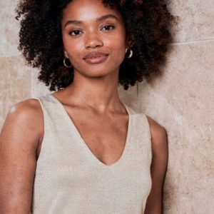 Une femme aux cheveux bouclés, portant un haut beige sans manches et des boucles d'oreilles, se tient debout contre un mur carrelé de couleur claire, en regardant la caméra.