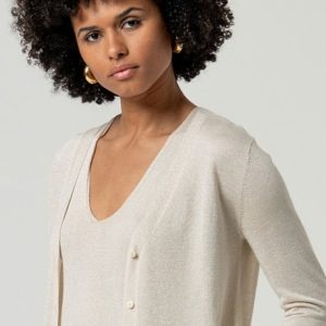 Une femme aux cheveux courts et bouclés porte un haut à col en V de couleur crème et un gilet assorti avec des boutons, debout sur un fond uni.