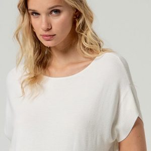 Une femme aux longs cheveux blonds, vêtue d'un ample haut blanc côtelé, se tient debout sur un fond clair uni, en regardant l'appareil photo.