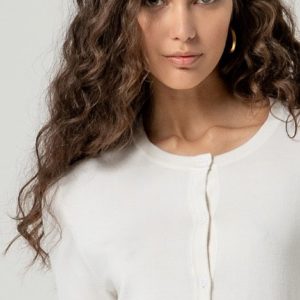 Femme aux longs cheveux bouclés portant un haut blanc boutonné et des boucles d'oreilles dorées, regardant l'appareil photo sur un fond uni.