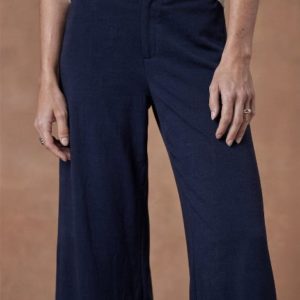 Personne portant un pantalon bleu marine à jambes larges et un haut blanc, les mains détendues sur les côtés, représentée de la taille à la mi-cuisse sur un fond brun uni.