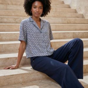 Une femme assise sur des marches en pierre, vêtue d'une chemise à manches courtes à motifs et d'un pantalon large de couleur marine, regarde l'appareil photo avec une expression neutre.