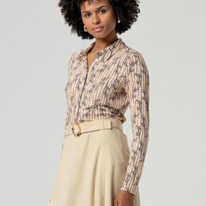 Femme portant un chemisier beige à motifs et à manches longues, rentré dans une jupe crème à taille haute avec une ceinture, debout sur un fond uni.