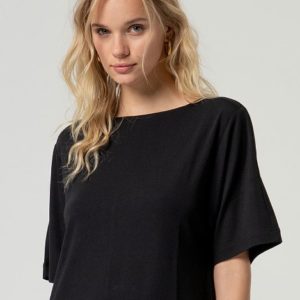 Une femme aux longs cheveux blonds, vêtue d'un haut noir ample, se tient debout sur un fond clair uni, face à la caméra.