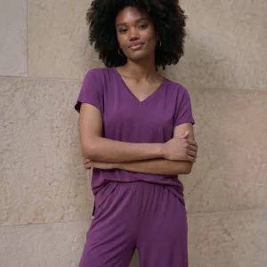 Une personne se tient contre un mur texturé beige, les bras croisés et le visage neutre, portant le T-shirt sans manches col en V Violet avec le pantalon assorti.