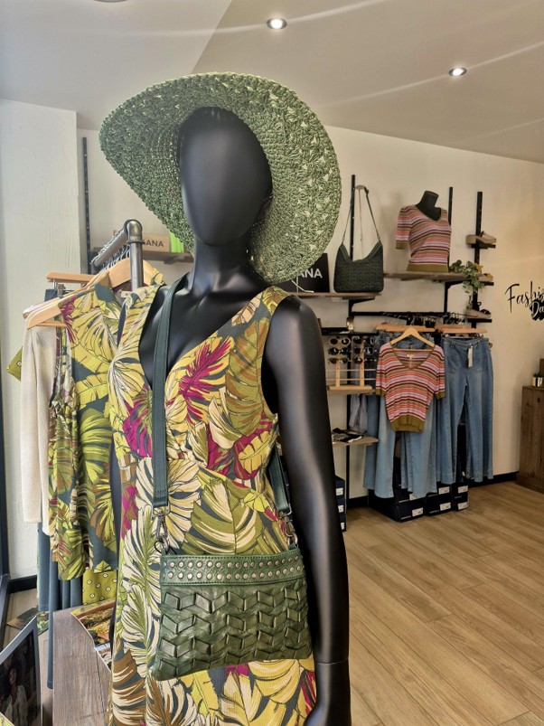 Mannequin portant un chapeau de paille vert, une combinaison à motifs floraux et un sac à main tressé vert, exposé dans une boutique de vêtements à Bergerac pendant les soldes d'été 2026, avec en arrière-plan des portants de chemises et de jeans.